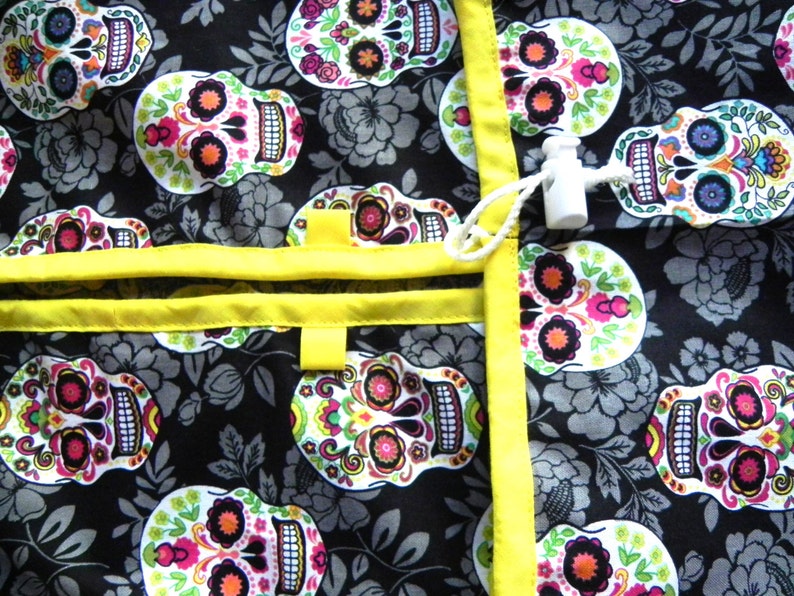 May include: A black fabric with a repeating pattern of colorful sugar skulls, flowers, and leaves. The fabric is trimmed with bright yellow ribbon and features a white toggle closure. The design is reminiscent of Dia de los Muertos.