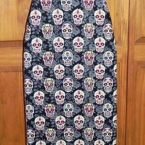 May include: An ironing board cover featuring a repeating pattern of colorful sugar skulls on a black background. The skulls have intricate floral and geometric designs in various colors. The cover is on a white ironing board frame.