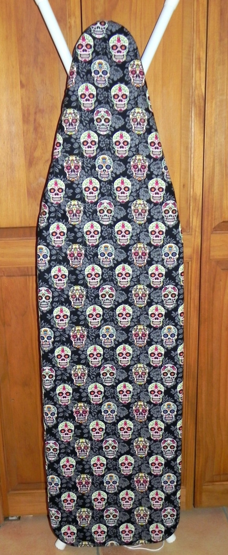 May include: An ironing board cover featuring a repeating pattern of colorful sugar skulls on a black background. The skulls have intricate floral and geometric designs in various colors. The cover is on a white ironing board frame.