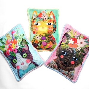 May include: Three rectangular cat-themed decorative pillows. Each pillow features a different cat face adorned with a floral crown. The pillows have a light blue, pink, and green background with a fringed edge.