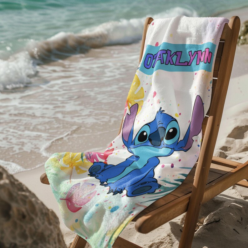 May include: A white beach towel featuring a blue cartoon character with large ears, colourful tropical leaf patterns in yellow and pink, and the name OAKLYNN printed in bold purple letters inside a light blue horizontal banner across the top.