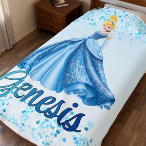 May include: A light blue fleece blanket featuring a graphic of a blonde character in a sparkling blue ballgown. The blanket is decorated with blue snowflake and diamond patterns and includes the name Genesis printed in large blue script lettering.