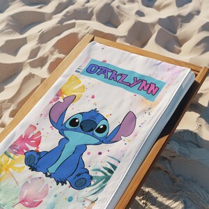 May include: A white rectangular beach towel featuring a blue cartoon character with large ears and a light blue chest patch. The towel displays the name OAKLYNN in pink letters on a blue banner, surrounded by colourful watercolour tropical leaves.
