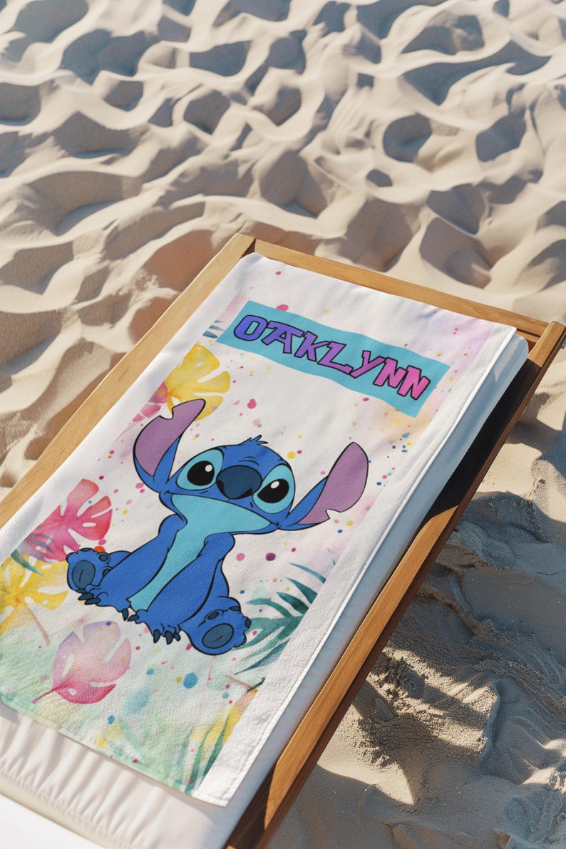 May include: A white rectangular beach towel featuring a blue cartoon character with large ears and a light blue chest patch. The towel displays the name OAKLYNN in pink letters on a blue banner, surrounded by colourful watercolour tropical leaves.