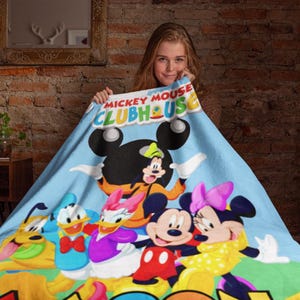 May include: A light blue fleece blanket featuring the Mickey Mouse Clubhouse logo at the top and a colorful graphic print of Mickey Mouse, Minnie Mouse, Donald Duck, Daisy Duck, Goofy, and Pluto standing together on a green grassy field.