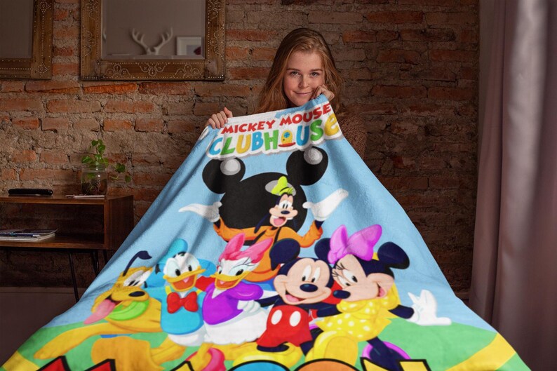 May include: A light blue fleece blanket featuring the Mickey Mouse Clubhouse logo at the top and a colorful graphic print of Mickey Mouse, Minnie Mouse, Donald Duck, Daisy Duck, Goofy, and Pluto standing together on a green grassy field.