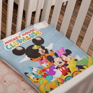 May include: A light blue blanket featuring the Mickey Mouse Clubhouse logo in red, yellow, and green text. The blanket displays a colorful graphic of Mickey Mouse, Minnie Mouse, Donald Duck, Daisy Duck, Goofy, and Pluto against a sky blue background.