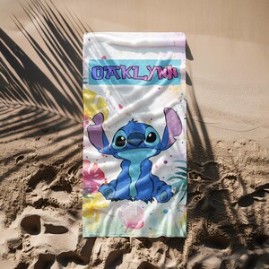 May include: A rectangular beach towel featuring a central illustration of the blue character Stitch. The name OAKLYNN is printed in a bold font within a light blue rectangular banner. The background includes colourful tropical leaf patterns.
