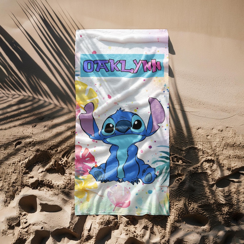 May include: A rectangular beach towel featuring a central illustration of the blue character Stitch. The name OAKLYNN is printed in a bold font within a light blue rectangular banner. The background includes colourful tropical leaf patterns.