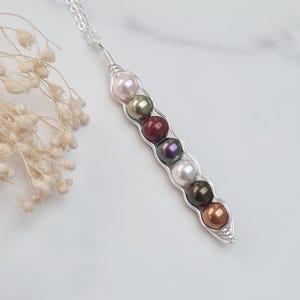 Puede incluir: Un collar de plata con un colgante de vaina de guisantes. El colgante, hecho de alambre de plata, contiene siete perlas en rosa, verde, rojo, morado, blanco, verde oscuro y bronce. El collar est&aacute; sobre una superficie blanca.