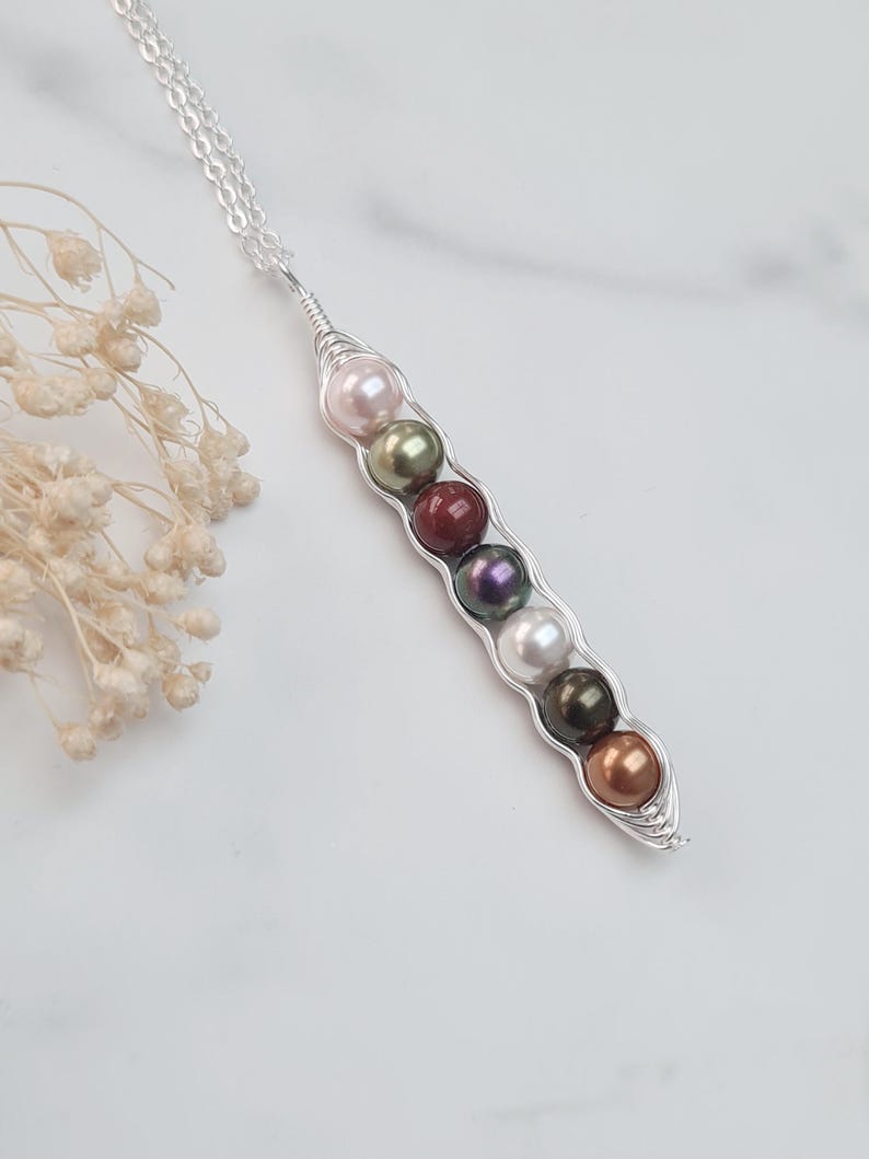 Puede incluir: Un collar de plata con un colgante de vaina de guisantes. El colgante, hecho de alambre de plata, contiene siete perlas en rosa, verde, rojo, morado, blanco, verde oscuro y bronce. El collar est&aacute; sobre una superficie blanca.