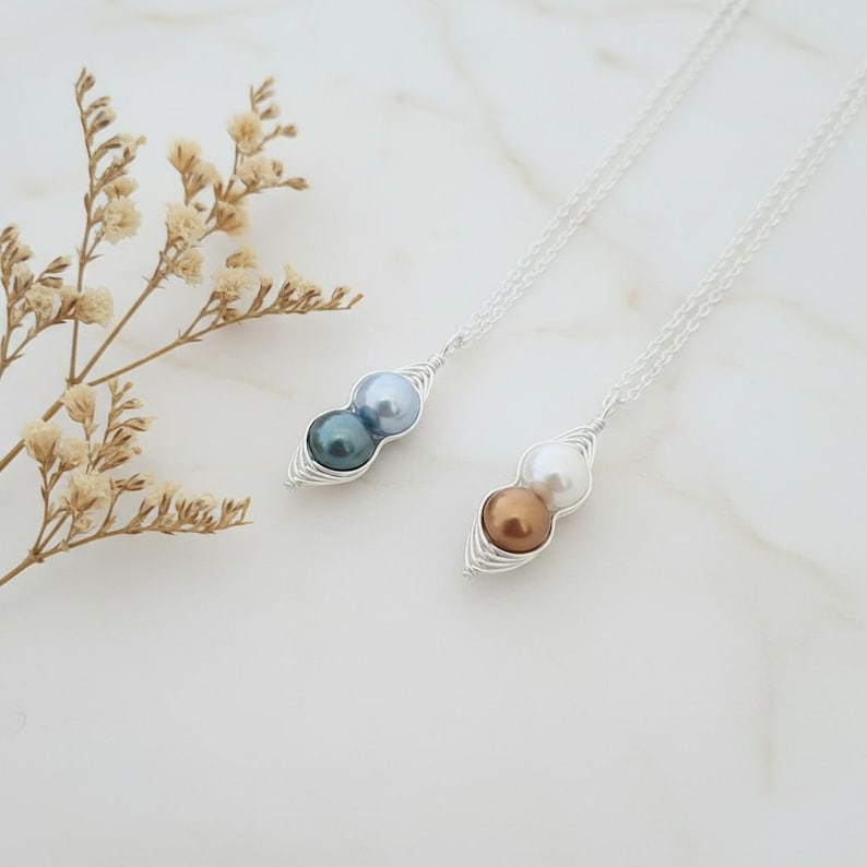 Puede incluir: Dos collares de plata, cada uno con un colgante en forma de vaina de guisantes. Uno contiene una perla azul oscuro y azul claro, el otro una perla blanca y bronce. Los collares est&aacute;n sobre una superficie blanca.