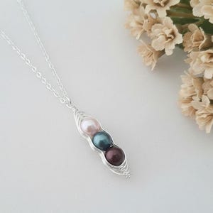 Puede incluir: Un collar de plata con un colgante de vaina de guisantes con tres perlas de colores: rosa, verde azulado y burdeos. El collar se muestra sobre una superficie blanca, con un ramo de flores beige en el fondo.