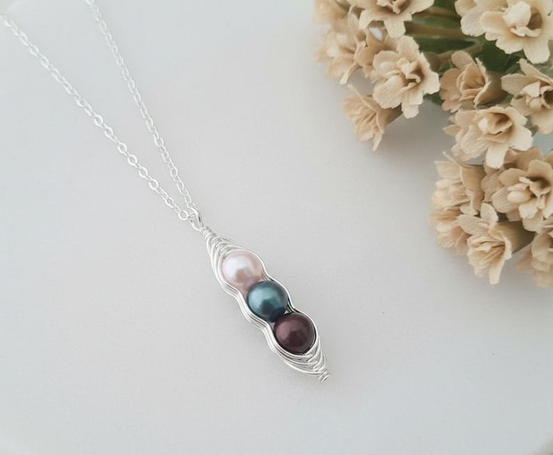 Puede incluir: Un collar de plata con un colgante de vaina de guisantes con tres perlas de colores: rosa, verde azulado y burdeos. El collar se muestra sobre una superficie blanca, con un ramo de flores beige en el fondo.