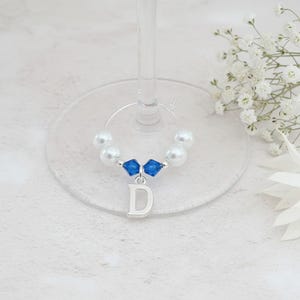 May include: A silver-toned wine glass charm featuring a circular wire hoop adorned with four white faux pearls and two faceted blue crystal beads. A silver-coloured metal letter D charm hangs from the centre of the hoop, creating a decorative accessory.