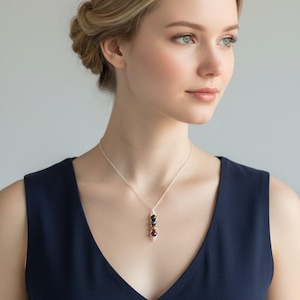 Puede incluir: Un collar de plata con un colgante con tres cuentas de color oscuro. Las cuentas est&aacute;n dispuestas verticalmente y son de diferentes tonos de marr&oacute;n y negro. La mujer de la imagen lleva un vestido azul marino.