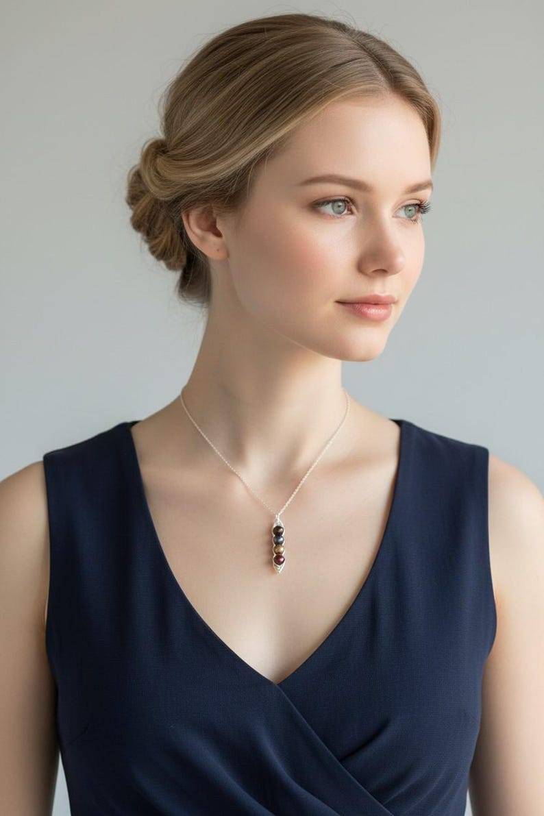 Puede incluir: Un collar de plata con un colgante con tres cuentas de color oscuro. Las cuentas est&aacute;n dispuestas verticalmente y son de diferentes tonos de marr&oacute;n y negro. La mujer de la imagen lleva un vestido azul marino.