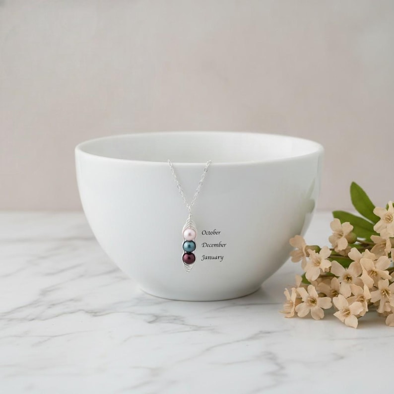 Puede incluir: Un collar de plata con tres cuentas de colores: rosa, celeste y rojo oscuro. El collar cuelga frente a un cuenco blanco. Las palabras "October, December, January" est&aacute;n impresas en el cuenco.