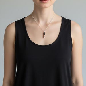 Puede incluir: Un collar de plata con un colgante con tres cuentas oscuras. Las cuentas est&aacute;n dispuestas verticalmente y suspendidas de una delicada cadena. El collar se lleva sobre una camiseta sin mangas negra.