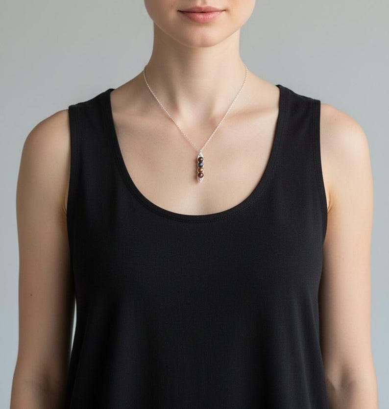 Puede incluir: Un collar de plata con un colgante con tres cuentas oscuras. Las cuentas est&aacute;n dispuestas verticalmente y suspendidas de una delicada cadena. El collar se lleva sobre una camiseta sin mangas negra.