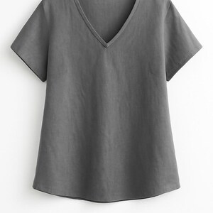Puede incluir: Una camiseta gris lisa con cuello en V y mangas cortas. La parte superior tiene un corte relajado y un diseño sencillo, adecuado para ropa informal. La tela parece suave y ligera.