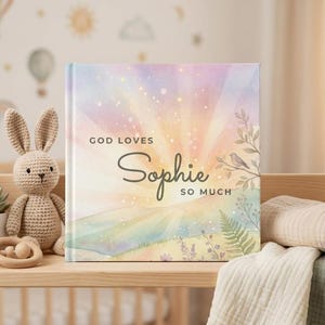 Puede incluir: Un libro infantil con el texto "God Loves Sophie So Much" en la portada, con un dise&ntilde;o de acuarela en colores pastel. Un conejito de ganchillo, una suculenta en maceta y un mordedor de madera est&aacute;n en una estanter&iacute;a de madera.