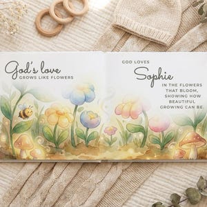 Puede incluir: Un libro abierto con el texto "God's love grows like flowers" y "God loves Sophie". Las ilustraciones presentan flores coloridas, una abeja y setas. El libro est&aacute; sobre una manta de punto beige con juguetes de madera.