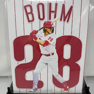 May include: A rectangular wall art print featuring a baseball player in a white pinstriped uniform with the number 28, holding a bat. The name BOHM is printed in red at the top, and a large red number 28 is centered behind the player on a white background.