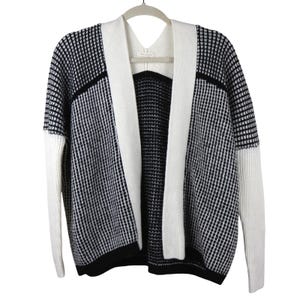 May include: A black and white open-front cardigan with a geometric pattern. The cardigan features white sleeves and a white shawl collar that frames the open front. The garment is hanging on a gray hanger.