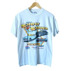 May include: White t-shirt featuring a colorful graphic for the 11th Annual Show 'N Shine car show. The design includes two classic cars, the show's name, and the location in Missouri. The shirt is hanging on a wooden hanger.