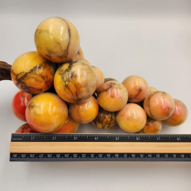 May include: A decorative cluster of marble grapes with a wooden stem. The grapes are various shades of yellow, orange, and red, with dark veining. A wooden ruler is placed next to the grapes, measuring approximately 10 inches.