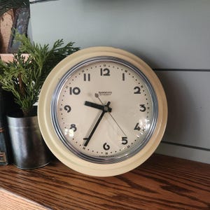 May include: A round wall clock with a cream-colored frame and a chrome bezel. The off-white clock face features black Arabic numerals, black hour and minute hands, a thin second hand, and the text Hammond Synchronous and Made in U.S.A.