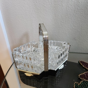 May include: A clear glass divided serving dish with a faceted, geometric pattern and a central vertical silver-toned metal handle. The dish features two distinct compartments with a starburst design at the base and a metallic frame at the bottom.