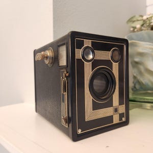 May include: A vintage black box camera featuring a textured leatherette finish and gold-toned geometric line accents on the front panel. The device includes a central lens, two circular viewfinders, a side winding knob, and a metal diaphragm lever.