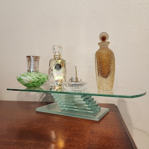 May include: A glass display shelf with a stacked base holds a green and white swirled glass lamp, a clear glass perfume bottle with gold accents, a small clear glass ring holder with a silver ring, and a tall amber textured glass bottle.