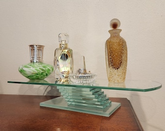 May include: A glass display shelf with a stacked base holds a green and white swirled glass lamp, a clear glass perfume bottle with gold accents, a small clear glass ring holder with a silver ring, and a tall amber textured glass bottle.