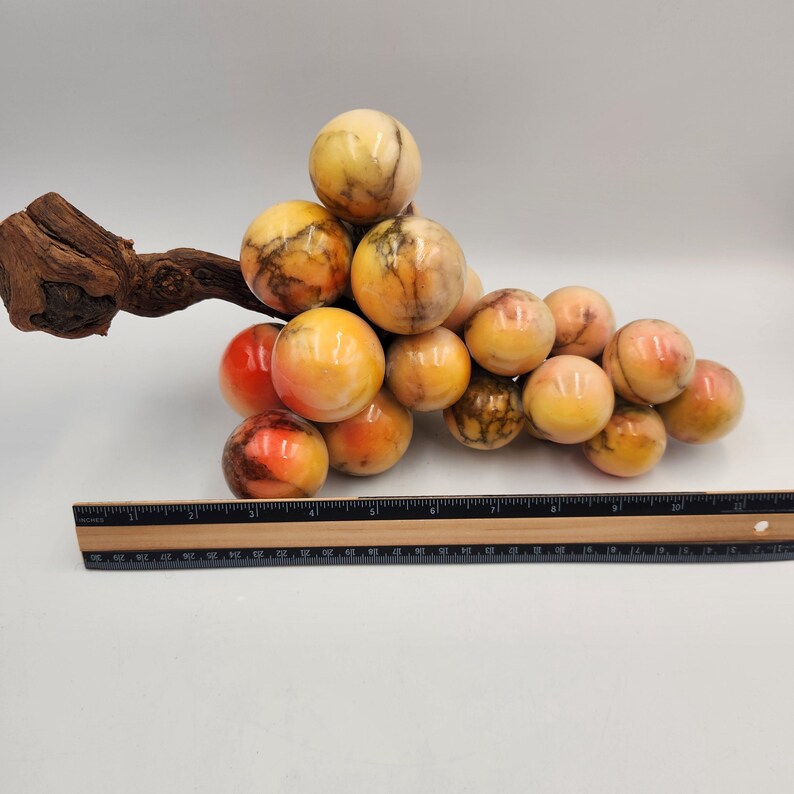 May include: A decorative cluster of marble grapes with a wooden stem. The grapes are a mix of yellow, orange, and red hues, with brown veining. A ruler is placed below the grapes for scale.