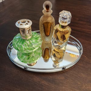 May include: A silver-rimmed mirrored tray holds three decorative glass bottles. One is green with white speckles, topped with a silver metal cap. Another is amber-colored with a brown stopper. The third is a faceted perfume bottle.