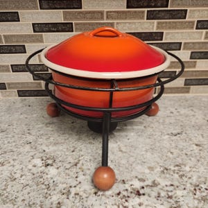 May include: An orange and red enameled fondue pot with a matching lid and a white rim. The pot sits in a black metal stand with wooden ball feet. The lid has a small handle.
