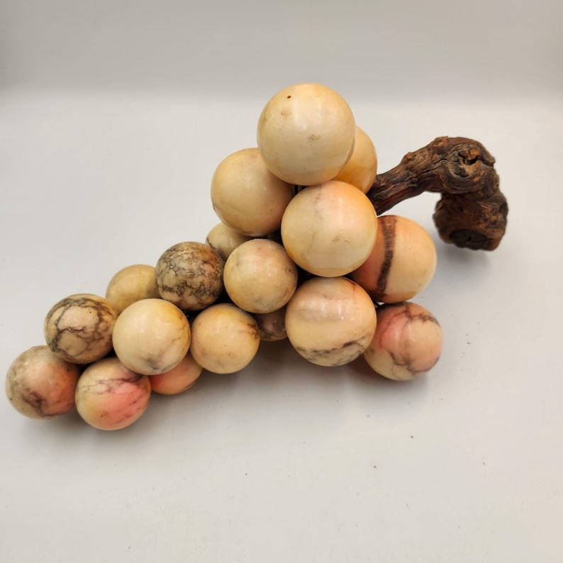 May include: A decorative sculpture of a bunch of grapes. The grapes are made of marble-like material in shades of cream, beige, and pink. The stem is a dark brown, textured piece of wood. The sculpture is a unique home decor item.