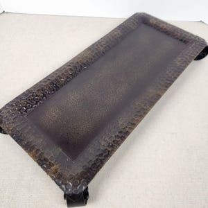May include: A rectangular metal tray with a dark brown finish and a hammered texture along the raised border. The center of the tray features a smooth, flat surface with a subtle speckled appearance. The tray is supported by four small, curved feet.