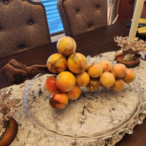 May include: A decorative cluster of marble grapes in shades of yellow, orange, and brown, resting on a clear glass plate. The grapes are attached to a natural wood branch. The arrangement sits on a patterned surface.
