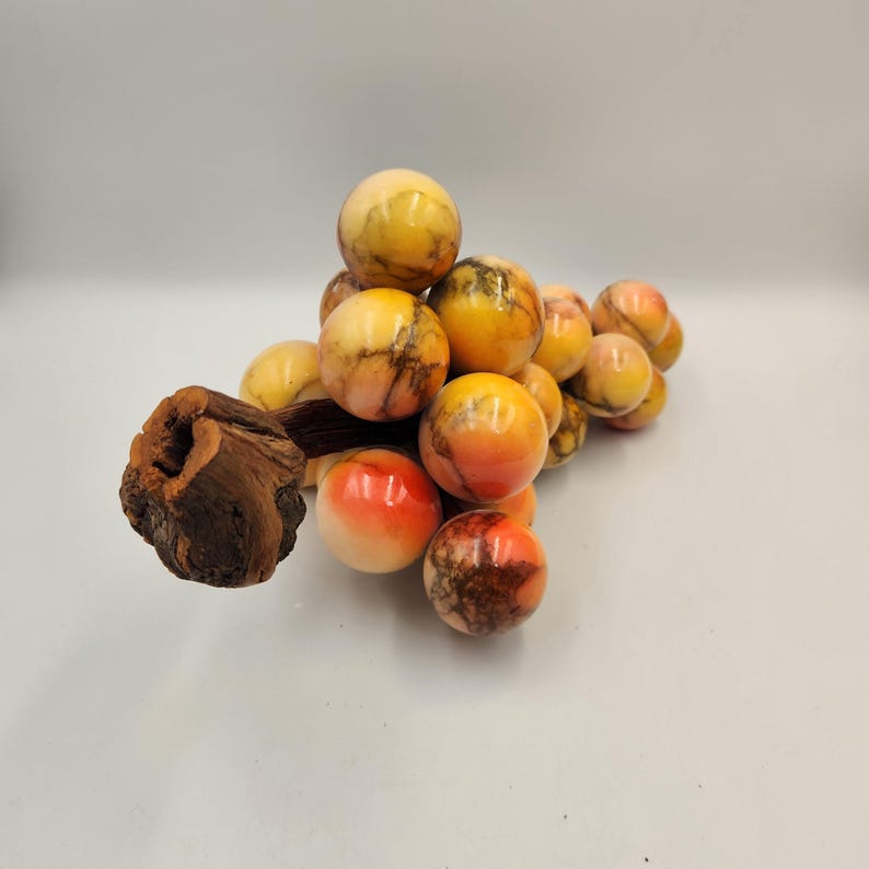 May include: A decorative cluster of marble spheres in shades of yellow, orange, and brown, resembling grapes. The spheres are attached to a piece of dark brown wood. The marble spheres have a polished, glossy finish.