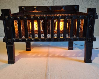 May include: A black metal electric fireplace insert featuring a rectangular frame with vertical bars, decorative pyramid studs, and a wire mesh backing. The unit is illuminated from within by two warm orange glowing light bulbs, creating a fire effect.