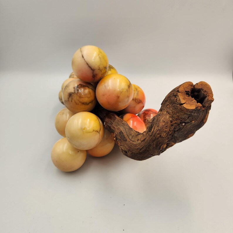 May include: A decorative cluster of marble spheres in shades of cream, yellow, and orange, resembling grapes. The spheres are attached to a piece of dark brown, textured wood, creating a unique and naturalistic sculpture. The background is a plain white.