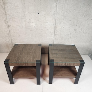 May include: Two square side tables with a wood grain finish and a lower shelf. The tables have black metal legs and a glass top. The tables are approximately 20 inches wide, 20 inches deep, and 20 inches tall.
