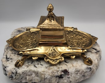 May include: A vintage brass inkwell with a central compartment and two side trays, resting on a stone base. The inkwell features ornate detailing and a decorative finial on top. The brass has a polished, golden finish.