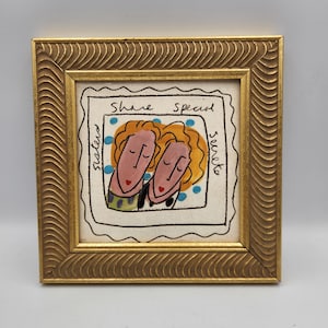 May include: A square framed artwork featuring a stylized illustration of two figures with orange hair and pink faces. The text "Sisters share special secrets" is written around the figures, which are set against a background of blue dots.
