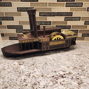 May include: A wooden model of a steamboat, featuring two smokestacks, a paddle wheel, and the name "ROBT. E. LEE" on the side. The model is primarily brown with gold accents and sits on a light-colored surface.