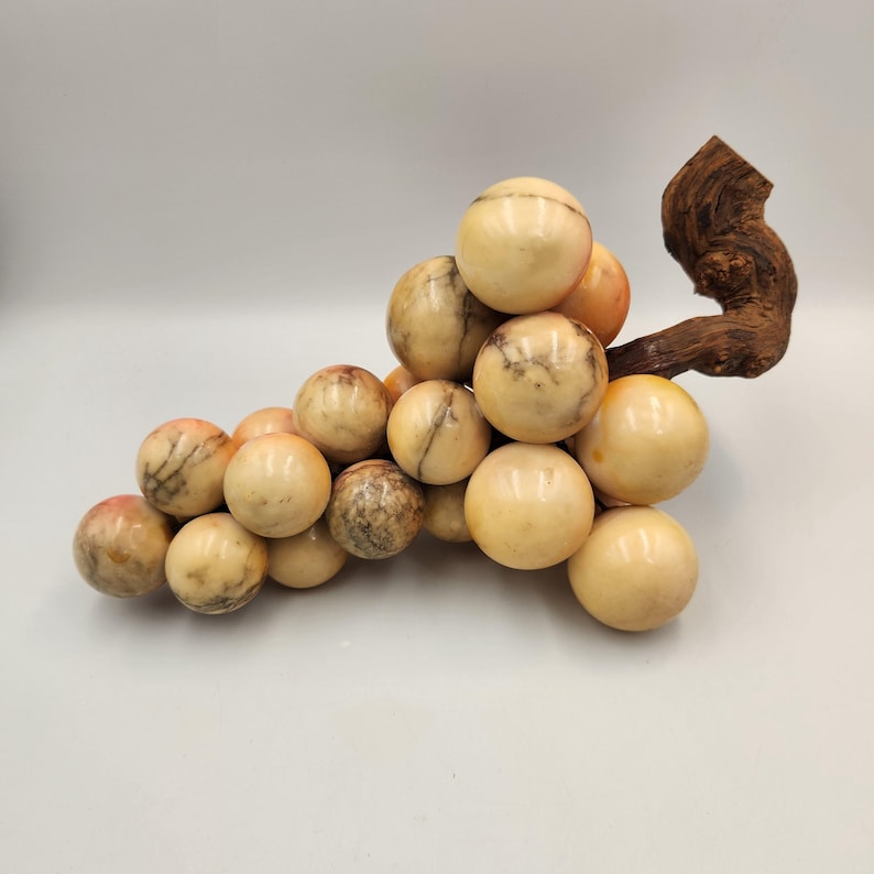 May include: A decorative sculpture of a bunch of grapes. The grapes are made of a creamy-colored stone with brown veining and a polished finish. The stem is a piece of dark, textured wood. A unique home decor item.
